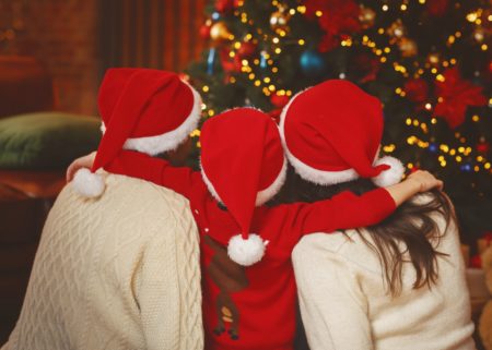 Small family of 3 with young child standing between parents with Santa hats on and looking at tree